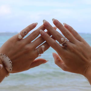 May include: A collection of silver rings adorned with pearls and seashells. The rings are displayed on a person's hands, with a seashell bracelet visible on the left wrist. The background features a blurred view of the ocean and sky.