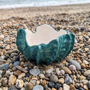 Könnte beinhalten: Eine türkisgrüne Keramikschale in Form einer Muschel mit glänzender Oberfläche. Der Innenraum ist cremeweiß. Die Schale liegt auf einem Bett aus Kieselsteinen und Sand, mit dem Meer im Hintergrund. Die strukturierte Außenseite der Schale ähnelt einer echten Muschel.