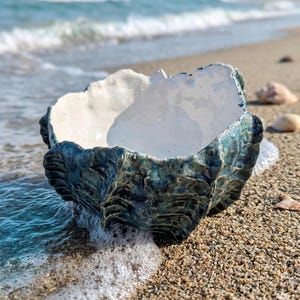 Könnte beinhalten: Eine Keramikschale mit einem strukturierten, muschelartigen Design. Die Schale ist hauptsächlich türkis und blau mit einem weißen Innenraum. Sie steht an einem sandigen Strand, an dessen Basis sanfte Wellen brechen und eine ruhige Küstenlandschaft schaffen.