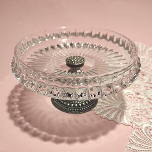 May include: A clear glass pedestal dish featuring a circular plate with a radial ribbed pattern and a scalloped edge. The dish is supported by a dark, metallic, textured base with a beaded design, providing a stable foundation for the glass top.