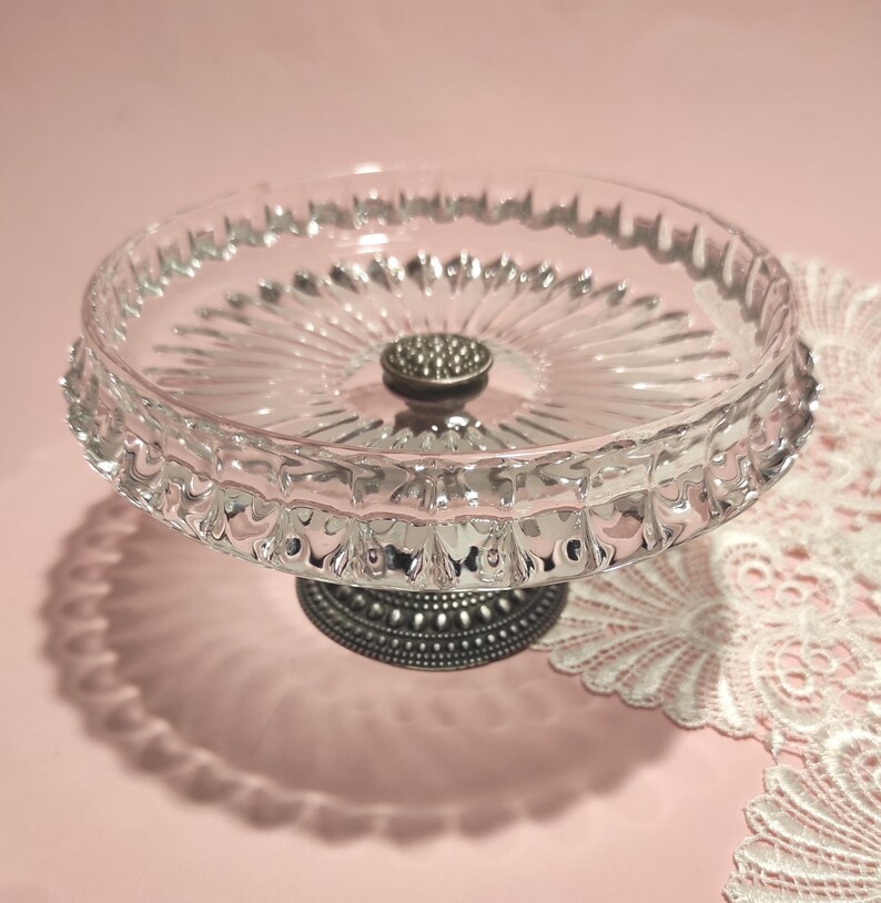 May include: A clear glass pedestal dish featuring a circular plate with a radial ribbed pattern and a scalloped edge. The dish is supported by a dark, metallic, textured base with a beaded design, providing a stable foundation for the glass top.