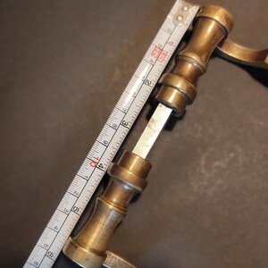 May include: A vintage brass door handle, approximately 4 inches long, with a decorative design. The handle is being measured with a white and silver measuring tape. The background is a dark, neutral color.