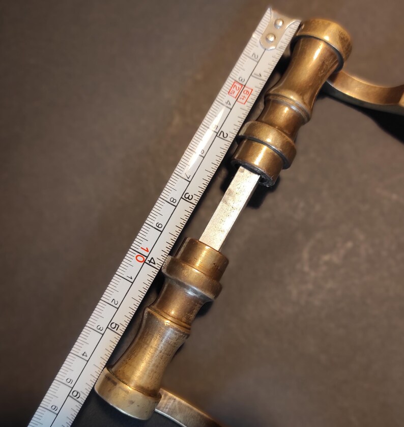 May include: A vintage brass door handle, approximately 4 inches long, with a decorative design. The handle is being measured with a white and silver measuring tape. The background is a dark, neutral color.