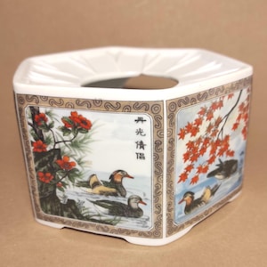 Puede incluir: Un calentador de té hexagonal de cerámica blanca con una abertura circular central. Los lados presentan ilustraciones pintadas de patos mandarines nadando, flores rojas y hojas de arce, con caracteres chinos negros impresos en un panel.