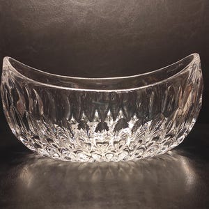 May include: A clear, decorative glass bowl with a crescent shape. The bowl has a textured, cut-glass design, creating a sparkling effect. The bowl is empty and sits on a dark surface.