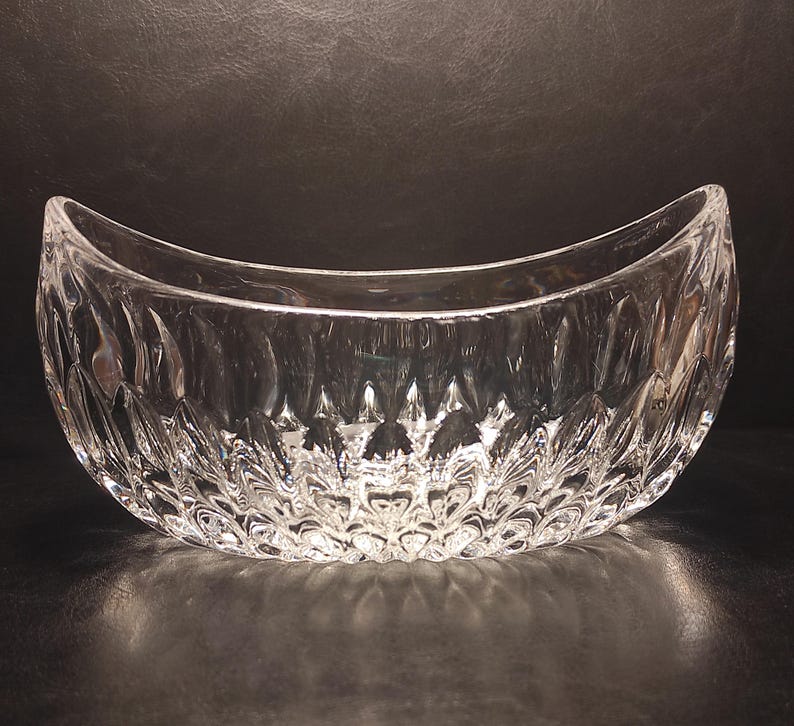 May include: A clear, decorative glass bowl with a crescent shape. The bowl has a textured, cut-glass design, creating a sparkling effect. The bowl is empty and sits on a dark surface.