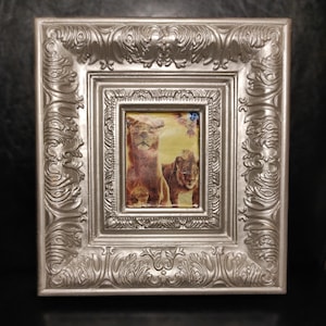 May include: A silver-colored, ornate framed picture featuring a print of two lions. The frame has a detailed, scrollwork design. The lions are depicted in shades of brown and tan against a yellow background.