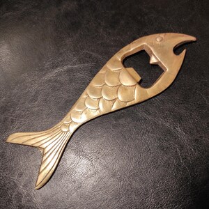 Könnte beinhalten: Ein messingfarbener Flaschenöffner in Form eines Fisches. Der Fisch hat ein detailliertes Schuppenmuster auf seinem Körper und einen flossenförmigen Schwanz. Der Flaschenöffner ist mit einem Mund und einem Flaschenöffner ausgestattet.