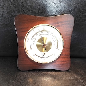 May include: A vintage barometer with a wooden frame and a circular dial. The dial features a brass center and a white face with black markings indicating pressure levels. The frame has a dark wood grain finish.