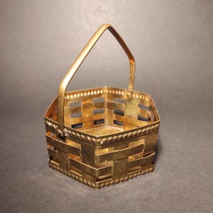 May include: A small, gold-coloured, hexagonal basket with a woven design and a handle. The basket is made of metal and has a decorative beaded trim.