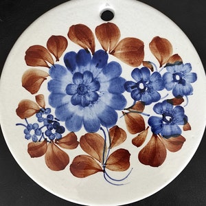 Puede incluir: Un plato de cerámica blanco con un diseño floral azul y marrón. El plato tiene un agujero en la parte superior para colgar.