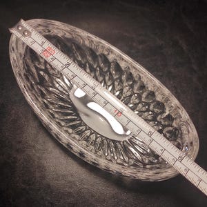 May include: A clear, oval-shaped crystal dish with a textured, diamond-cut pattern. A silver spoon rests inside the dish. The dish is approximately 7 inches long, as measured by a ruler.