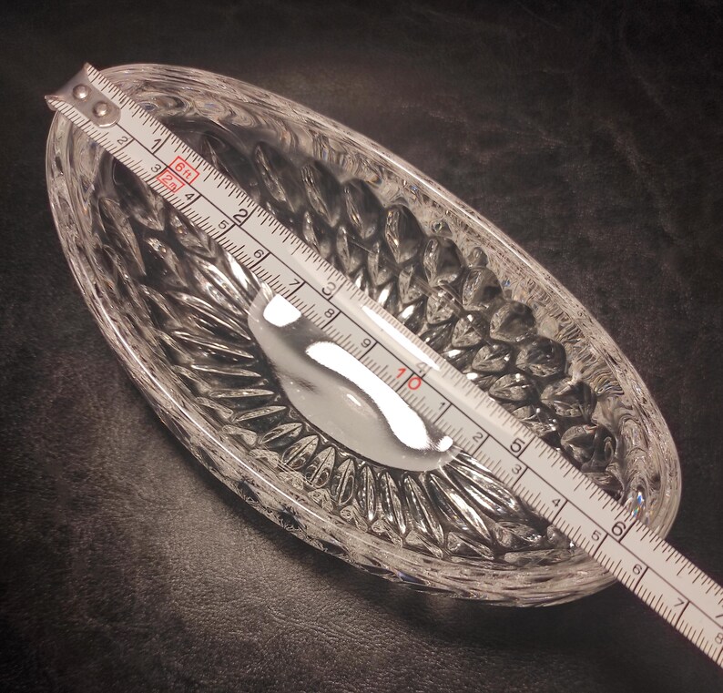 May include: A clear, oval-shaped crystal dish with a textured, diamond-cut pattern. A silver spoon rests inside the dish. The dish is approximately 7 inches long, as measured by a ruler.