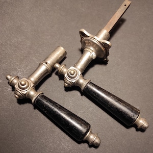 May include: Two antique door handles with a silver finish and black wooden grips. The handles have ornate detailing with decorative metal accents. The handles are likely vintage or antique, and are designed for a door.