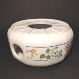 Op de afbeelding: Witte keramische theewarmhouder met een bloemenontwerp met oranje, blauwe en roze bloemen. De warmer heeft een cirkelvormige opening aan de bovenkant en een handgreepachtige opening aan de zijkant. Een blauwe streep accentueert het bloemenpatroon.