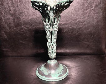 Puede incluir: Un candelabero de metal con acabado en cardenillo que presenta una pátina desgastada en tonos verdes y cobrizos. El tallo tiene un diseño intrincado de filigrana calada que se ensancha hacia arriba, sobre una base circular lisa.