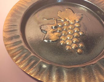 May include: A round copper-toned metal plate featuring a central embossed design of a cluster of grapes with leaves. The plate has a dark, reflective finish in the center and a wide, textured, fluted rim with a lighter metallic copper sheen.