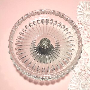 May include: A clear glass dish with a scalloped rim and a radial pattern of embossed ridges extending from the centre. A circular, metallic silver-toned handle with a textured, dotted surface is positioned in the middle of the glass bowl.