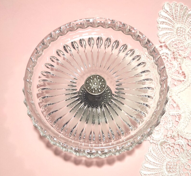 May include: A clear glass dish with a scalloped rim and a radial pattern of embossed ridges extending from the centre. A circular, metallic silver-toned handle with a textured, dotted surface is positioned in the middle of the glass bowl.