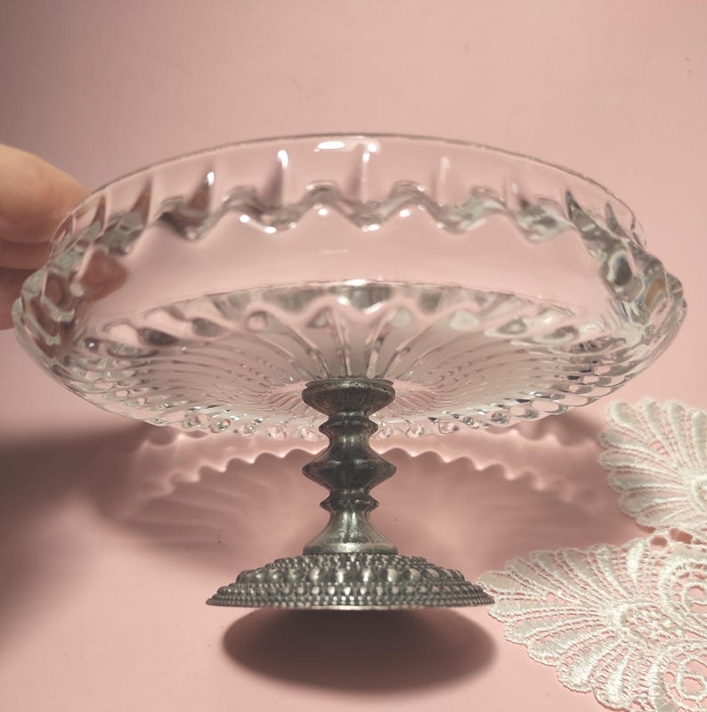 May include: A clear glass pedestal dish featuring a scalloped rim and a radiating sunburst pattern on the base. The bowl is supported by a dark, textured metal stand with a wide, circular base that has a fine, beaded edge detail throughout.