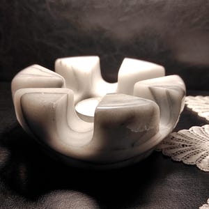 May include: A white marble candle holder with grey veining, featuring a circular base and five distinct, curved, wedge-shaped segments that rise upward to create a central opening designed to hold a small tea light candle in the middle.