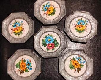 Puede incluir: Seis platos decorativos octogonales con un marco de metal plateado. Cada plato presenta un diseño floral pintado a mano en colores vibrantes como amarillo, azul y rojo sobre un fondo blanco. Los platos están dispuestos sobre una superficie oscura.