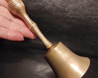 May include: A vintage brass bell with a rounded handle and a flared base. The handle has a decorative ball at the top with horizontal ridges. The bell is a warm, golden color and appears to be made of metal.