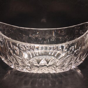 May include: A clear, oval-shaped glass bowl with a detailed, textured pattern. The bowl has a wide, curved opening and sits on a dark surface. The intricate design catches the light, creating a sparkling effect. This decorative bowl is suitable for serving or display.