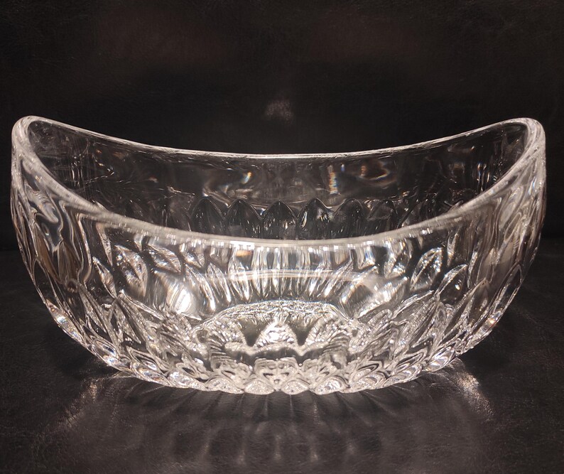 May include: A clear, oval-shaped glass bowl with a detailed, textured pattern. The bowl has a wide, curved opening and sits on a dark surface. The intricate design catches the light, creating a sparkling effect. This decorative bowl is suitable for serving or display.