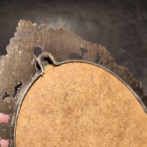 May include: The back of a round, ornate, gold-toned picture frame with a brown cardboard backing. The frame has a decorative, scalloped edge.