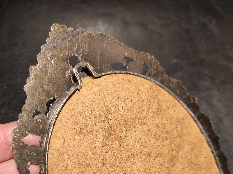 May include: The back of a round, ornate, gold-toned picture frame with a brown cardboard backing. The frame has a decorative, scalloped edge.