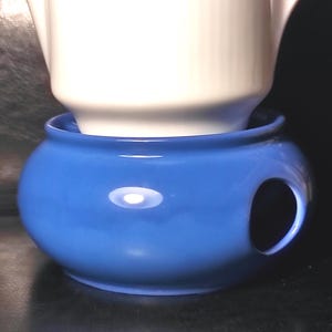 Pode incluir: Um aquecedor de chá de cerâmica azul cobalto com uma chaleira branca dentro. O aquecedor tem uma forma arredondada com uma abertura circular para uma vela. A chaleira é branca.
