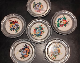 Puede incluir: Juego de seis posavasos decorativos. Cada posavasos tiene un borde de metal plateado y un diseño floral. El centro de cada posavasos presenta una disposición diferente de flores coloridas sobre un fondo blanco agrietado. Sobre una superficie oscura.