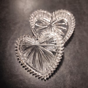 May include: Two clear, heart-shaped glass trinket boxes. The boxes have a starburst design in the center and a scalloped edge. The boxes are on a dark surface.