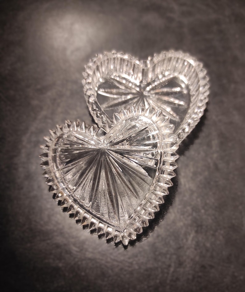 May include: Two clear, heart-shaped glass trinket boxes. The boxes have a starburst design in the center and a scalloped edge. The boxes are on a dark surface.