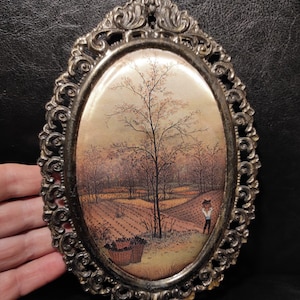May include: An oval-shaped framed print with a vintage-style image of a rural landscape with a tree and a person in the distance. The frame is ornate and has a gold finish.
