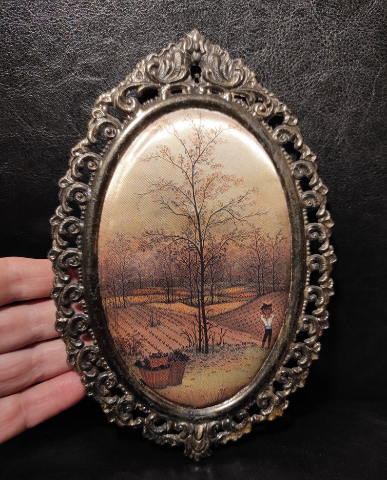 May include: An oval-shaped framed print with a vintage-style image of a rural landscape with a tree and a person in the distance. The frame is ornate and has a gold finish.