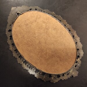 May include: An oval-shaped, silver-toned picture frame with a decorative, ornate border. The frame is empty and has a brown backing.