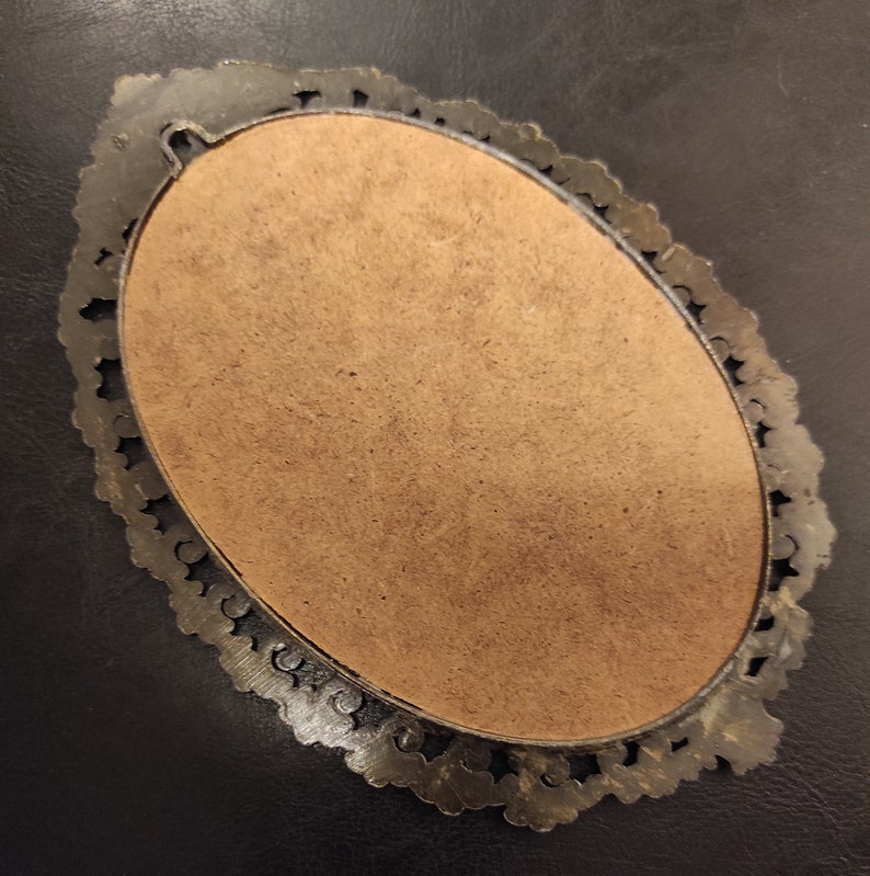 May include: An oval-shaped, silver-toned picture frame with a decorative, ornate border. The frame is empty and has a brown backing.