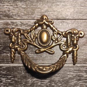 May include: Ornate gold-toned metal drawer pull with a decorative design featuring two stylised animal heads, a central oval, and a floral motif.