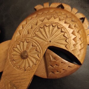 Op de afbeelding: Een houten notenkraker gesneden met ingewikkelde details. De notenkraker is gevormd als een helm met een cirkelvormig ontwerp aan de voorkant en een waaiervormig ontwerp aan de bovenkant.