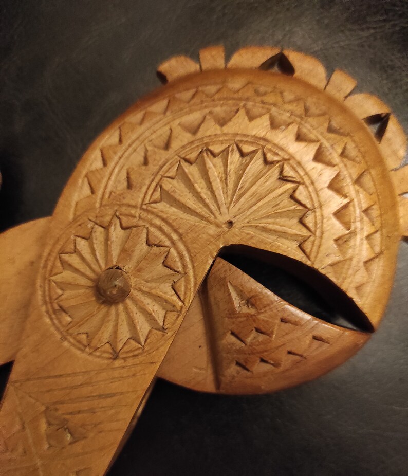 Op de afbeelding: Een houten notenkraker gesneden met ingewikkelde details. De notenkraker is gevormd als een helm met een cirkelvormig ontwerp aan de voorkant en een waaiervormig ontwerp aan de bovenkant.
