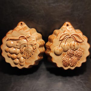 May include: Two ceramic moulds with a scalloped edge. One mould features a grape design, the other cherries and raspberries. Both are tan with brown detailing and a hole for hanging.