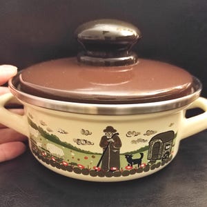 Puede incluir: Una pequeña olla de esmalte color crema con tapa y asa marrones. La olla presenta una escena pastoral con una figura, ovejas y una caravana. La tapa tiene un pomo marrón oscuro. La olla tiene un borde plateado.