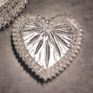 May include: A clear, heart-shaped trinket dish with a starburst design radiating from the center. The dish has a scalloped edge and is made of cut crystal. The image shows the dish reflecting light.