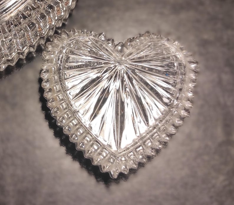 May include: A clear, heart-shaped trinket dish with a starburst design radiating from the center. The dish has a scalloped edge and is made of cut crystal. The image shows the dish reflecting light.