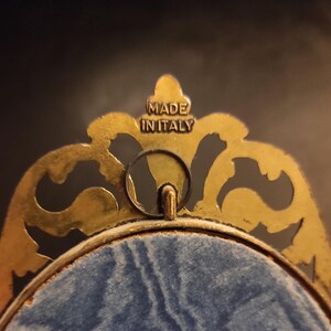 Puede incluir: Primer plano de un marco decorativo dorado con adornos ornamentados. El marco tiene un respaldo de terciopelo azul y un peque&ntilde;o lazo para colgar. El texto "MADE IN ITALY" es visible en la parte superior del marco.