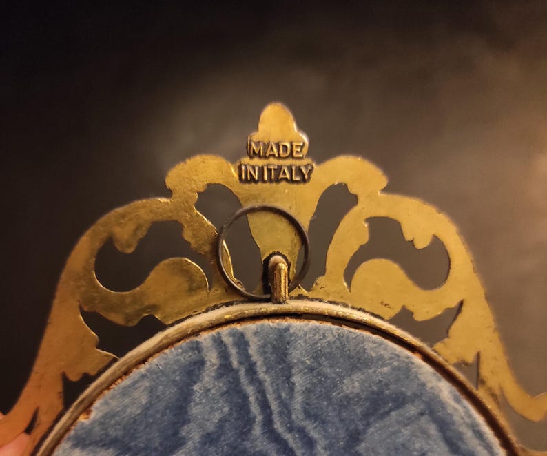 Puede incluir: Primer plano de un marco decorativo dorado con adornos ornamentados. El marco tiene un respaldo de terciopelo azul y un peque&ntilde;o lazo para colgar. El texto "MADE IN ITALY" es visible en la parte superior del marco.