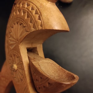 Op de afbeelding: Een houten lepel met ingewikkelde houtsnijwerk, met een bloemenmotief en een geometrisch patroon. De lepel is bevestigd aan een grotere houten structuur, ook gesneden met een bloemenmotief.