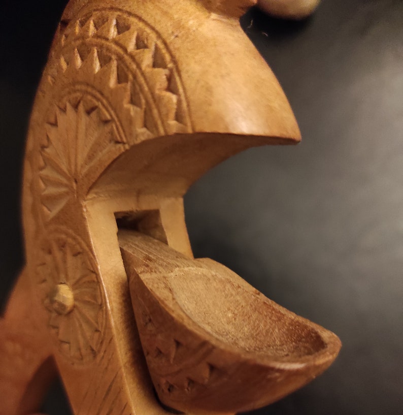 Op de afbeelding: Een houten lepel met ingewikkelde houtsnijwerk, met een bloemenmotief en een geometrisch patroon. De lepel is bevestigd aan een grotere houten structuur, ook gesneden met een bloemenmotief.